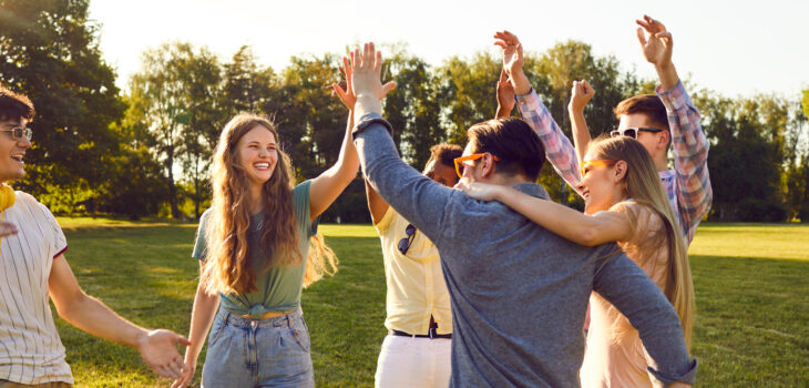 Bunch of happy young friends all together having fun on warm sunny day in summer park. Diverse group of cheerful joyful positive people standing on green lawn, smiling and giving each other high five